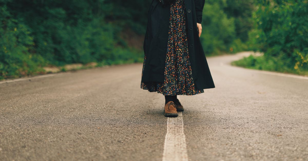 Woman in a trench coat and floral skirt walking on a tranquil countryside road.