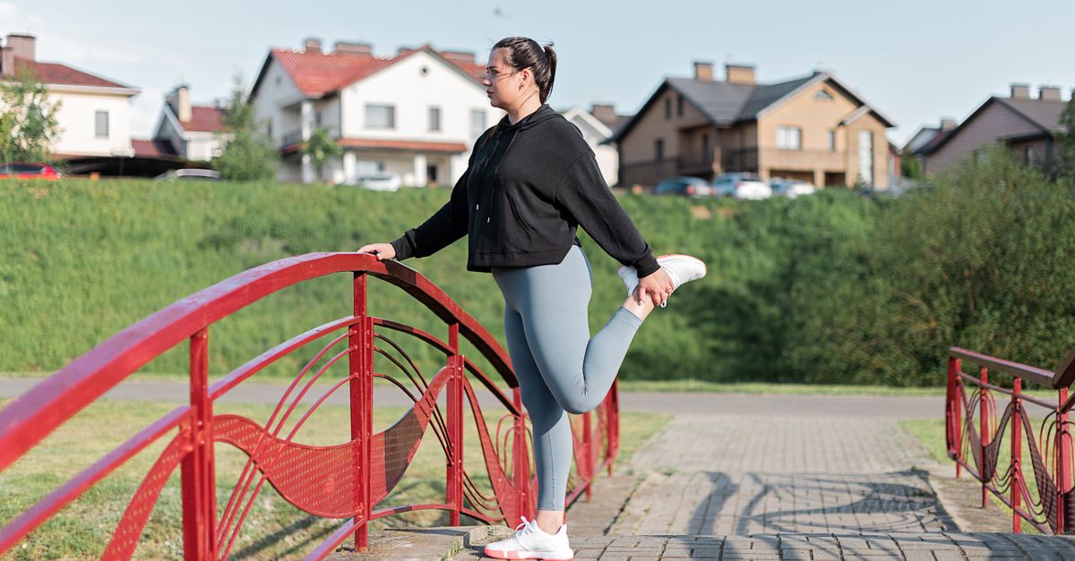 A woman in active wear performs a stretching exercise on a red bridge in a suburban park setting.