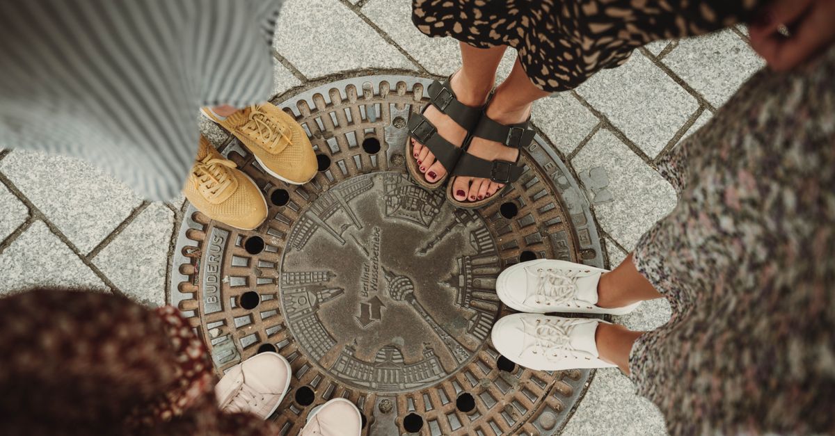 Top view of friends' shoes and sandals around a Berlin manhole cover, symbolizing unity.