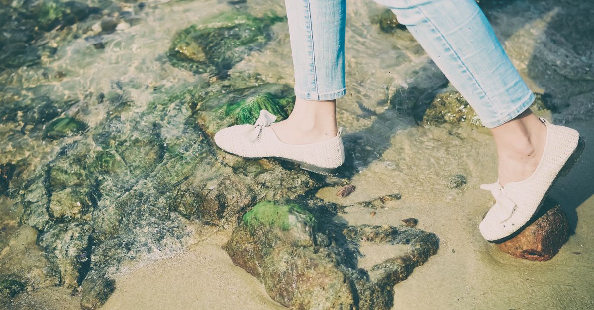 Person in casual shoes walking on rocky beach shore with clear water.