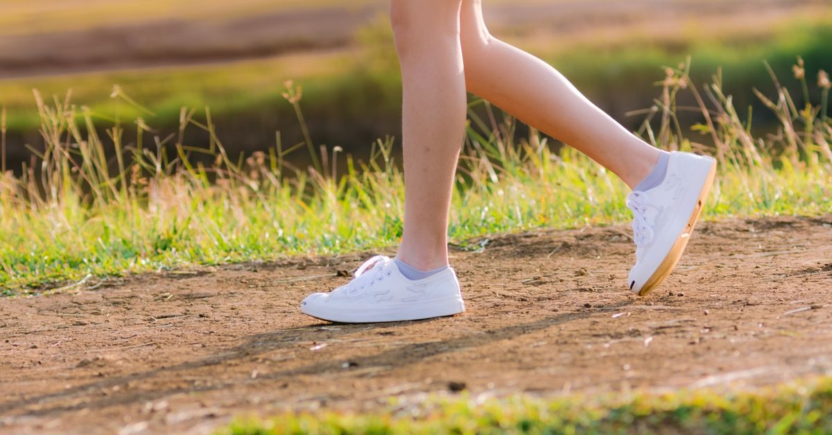 A woman walks on a dirt path in a sunlit park, wearing white sneakers and a skirt.