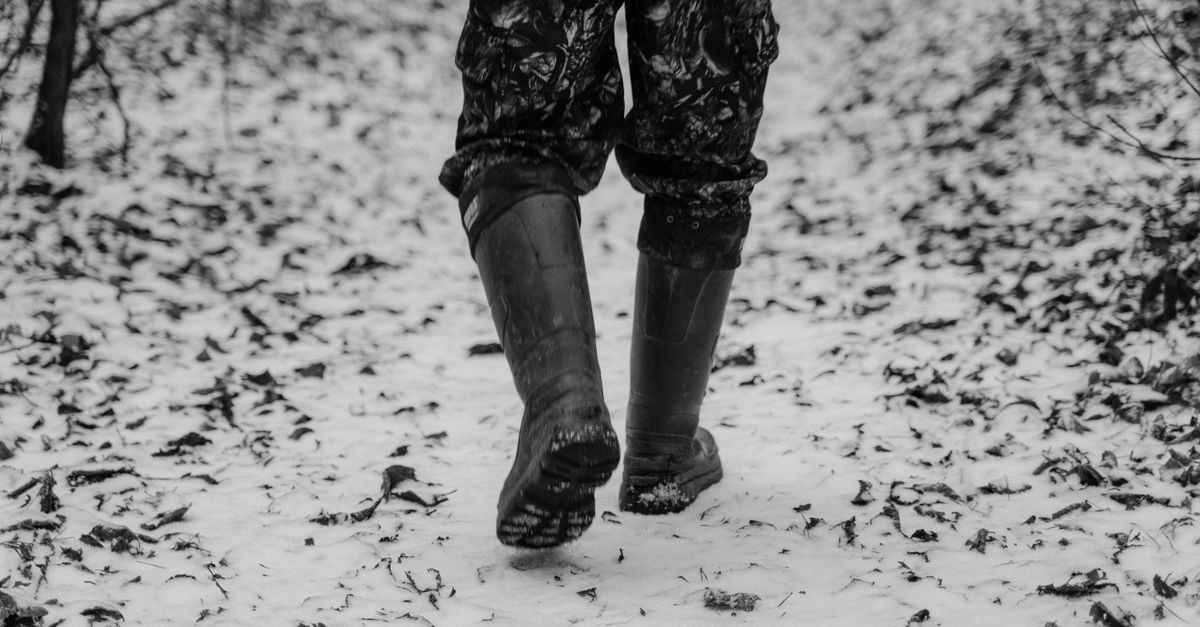 Person walking in snow with boots on a wintery forest trail captured in black and white.