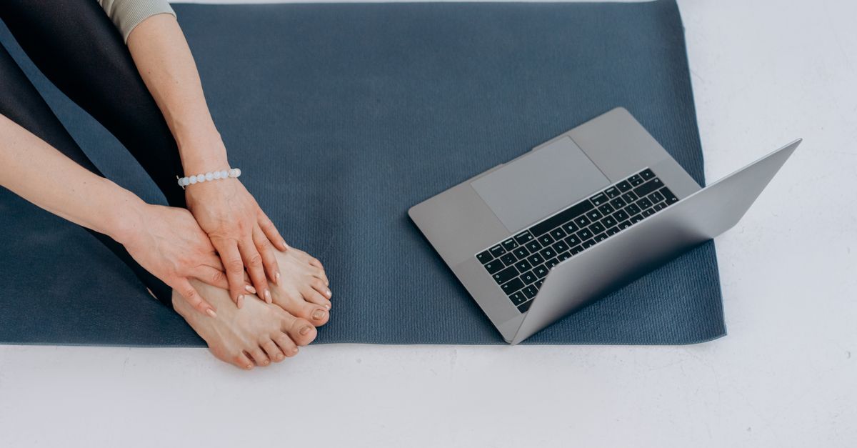 Individual on yoga mat practicing stretches alongside a laptop for online fitness classes.