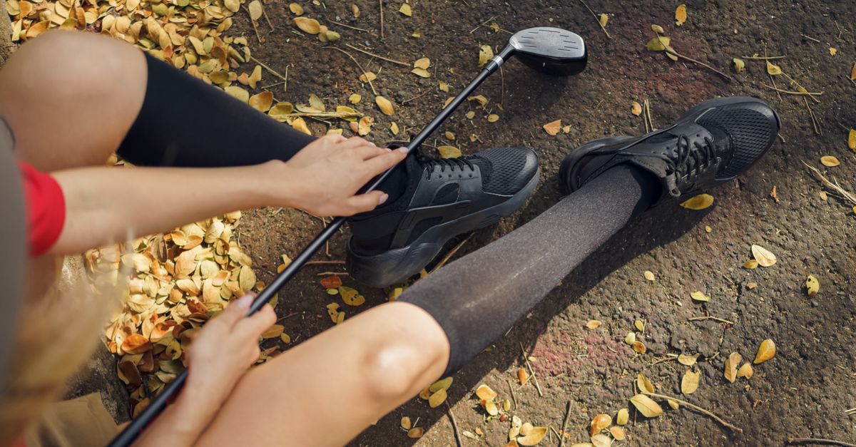 A person holding a golf club sits on the ground surrounded by autumn leaves, wearing black shoes.