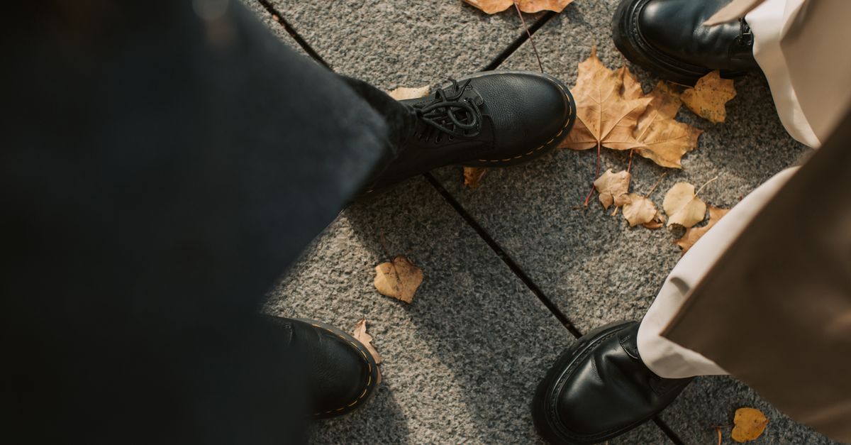 Overhead view of two people in leather shoes standing on a leaf-strewn pavement in fall.