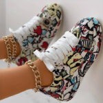Une paire de pieds portant des baskets colorées à motifs graffitis avec des lacets blancs. Les baskets ont des motifs audacieux et artistiques en rouge, bleu, noir et jaune. Chaque cheville est ornée d'une chaîne de cheville épaisse en or.