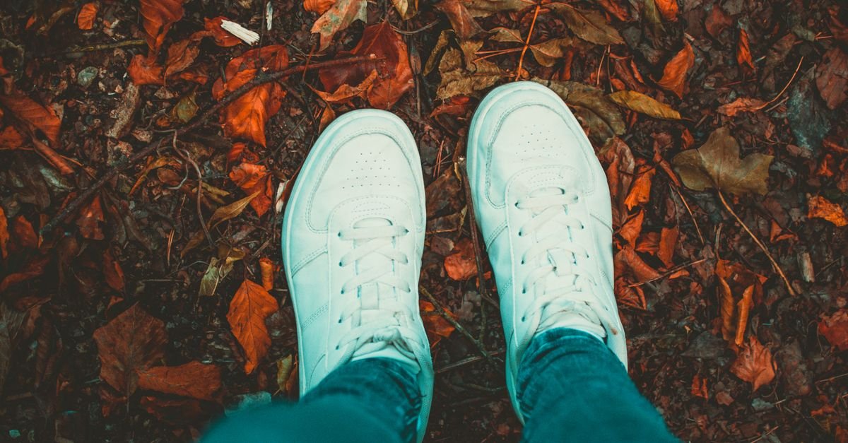 Person wearing white sneakers standing on a path covered with autumn leaves, showcasing a fall vibe.