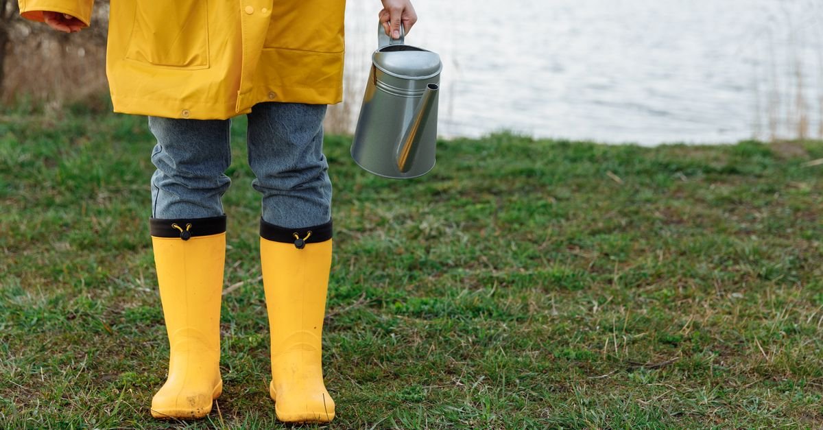 Person wearing yellow rain gear and holding a watering can by the lake.